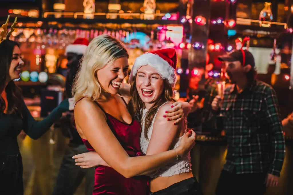 Two women wearing festive clothing, including Santa hats, hug and laugh in a lively bar setting with holiday lights in the background.